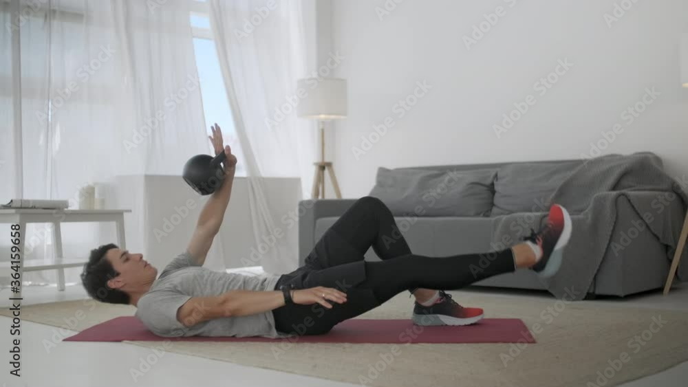Handsome Young Man in a Gray T-Shirt and Black Shorts Does Sports at Home Online. Strong Athletic Male is Doing Crisscross Crunch Exercises at Home in His Spacious and Sunny Living Room. Handsome Young Man in a Gray T-Shirt and Black Shorts Does Sports at Home Online. Strong Athletic Male is Doing Crisscross Crunch Exercises at Home in His Spacious and Sunny Living Room.