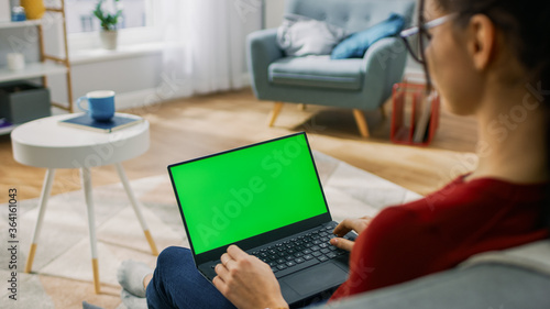 Young Woman at Home Works on a Laptop Computer with Green Mock-up Screen. She s Sitting On a Couch in His Cozy Living Room. Over the Shoulder Shot