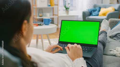 Woman at Home Lying on a Couch Working on Laptop Computer with Green Mock-up Screen. Girl Using Computer for Internet and Social Networks Browsing. Over the Shoulder Shot.