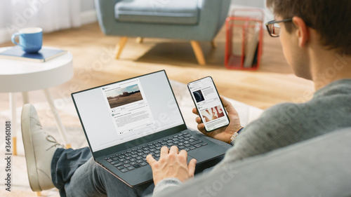 Young Man at Home is Using Laptop Computer, Srufing the Net, Watching News, while Holding Smartphone with Social Media Interface. He's Sitting On a Couch in His Cozy Living Room. Over Shoulder Shot