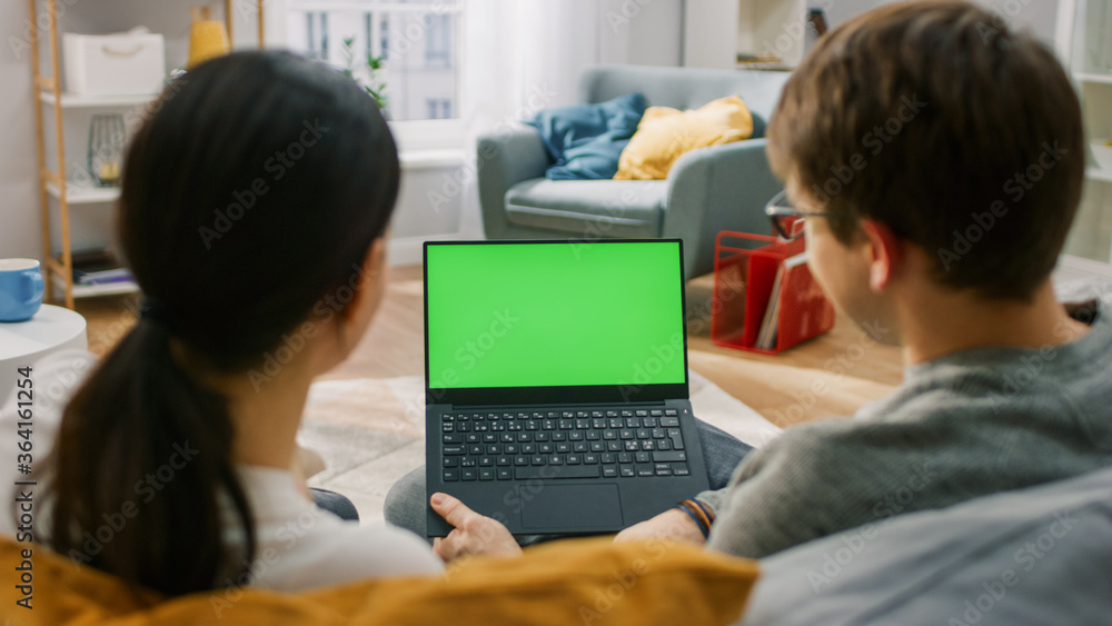 Young Man and Woman at Home Using Green Mock-up Screen Laptop Computer While Sitting on Couch in Living Room. Couple in Love Talking and Watching TV Programme. Back View Shot. Young Man and Woman at Home Using Green Mock-up Screen Laptop Computer While Sitting on Couch in Living Room. Couple in Love Talking and Watching TV Programme. Back View Shot.