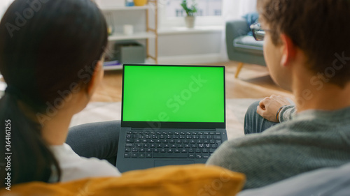 Young Man and Woman at Home Using Green Mock-up Screen Laptop Computer While Sitting on Couch in Living Room. Couple in Love Talking and Watching TV Programme. Back View Camera Shot.