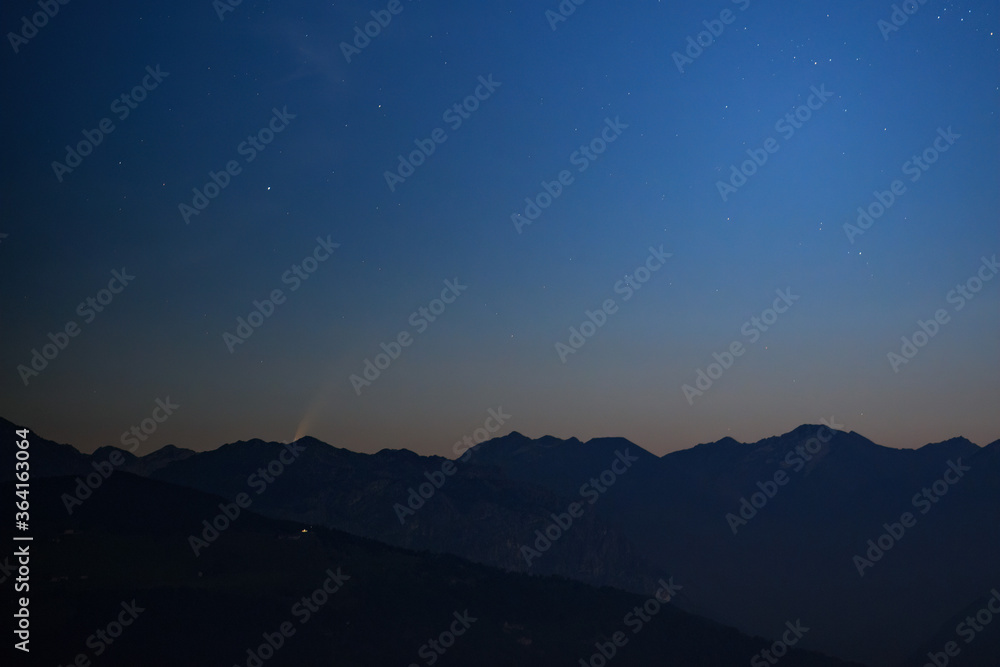 Paesaggio montano alle prime luci dell’alba con nel cielo la cometa C2020 F3 NEOWISE che spunta da dietro le cime dei monti.