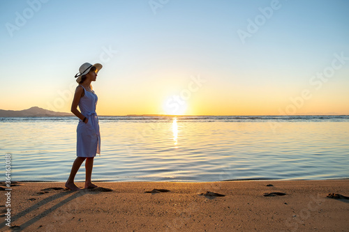 Young woman in straw hat and a dress walking alone on empty sand beach at sunset sea shore. Lonely girl looking at horizon over calm ocean surface on vacation trip.