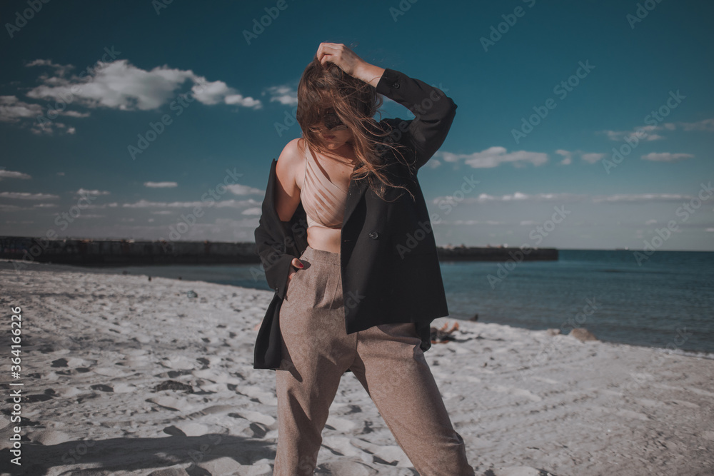 young brunette with long hair in a dark jacket and trousers in sunglasses on the seashore