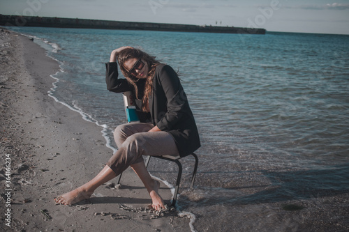 young brunette with long hair in a dark jacket and trousers in sunglasses with a chair on the seashore