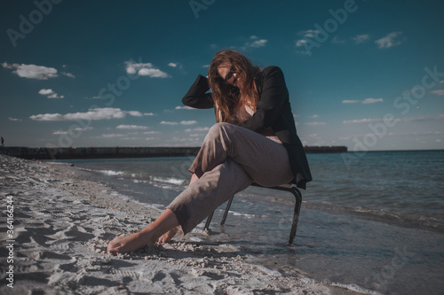 young brunette with long hair in a dark jacket and trousers in sunglasses with a chair on the seashore