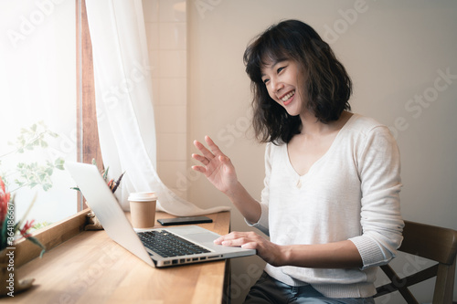 Young Asian woman working and video conferencing with laptop computer. A happy woman with smiley face working from home. A cup of coffee on wooden table. Work from home concept