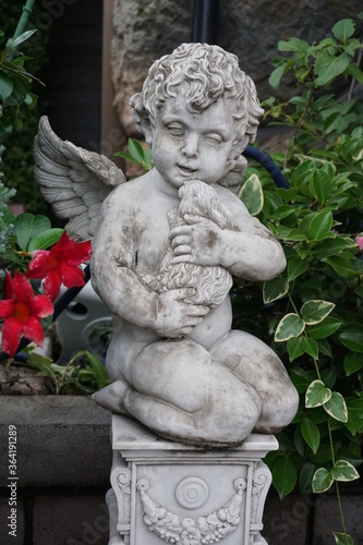 Statue of a cupid, angel holding a doll in a flowered garden 1
花の咲いた庭の中にある人形を抱えたキューピッド、天使の像 1