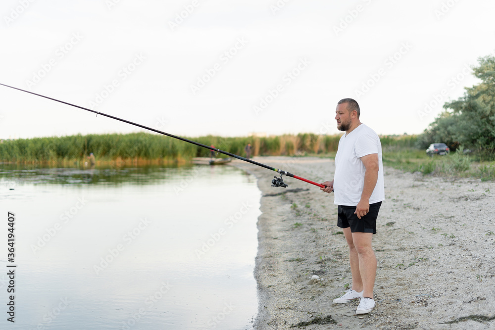 A man fishing. In summer, a dressed man with a fishing rod in his hands. Fishing on the shore of a pond. A middle-aged man is waiting for a bite. He is concentrated A man fishing. In summer, a dressed man with a fishing rod in his hands. Fishing on the shore of a pond. A middle-aged man is waiting for a bite. He is concentrated