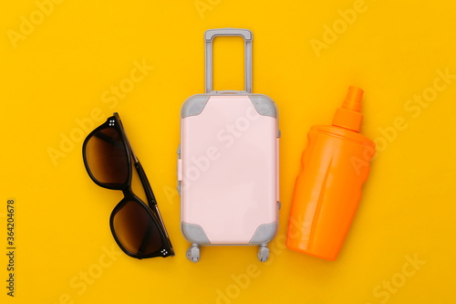 Beach vacation, travel concept. Mini toy travel luggage and sunblock bottle, sunglasses on yellow background. Top view. Flat lay
