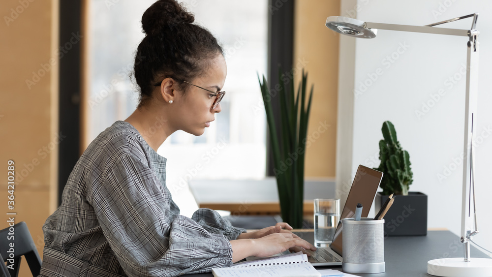Focused biracial young woman in glasses sit at desk look at laptop screen work consulting client online, serious ethnic female employee type on computer, prepare business report or project in office Focused biracial young woman in glasses sit at desk look at laptop screen work consulting client online, serious ethnic female employee type on computer, prepare business report or project in office