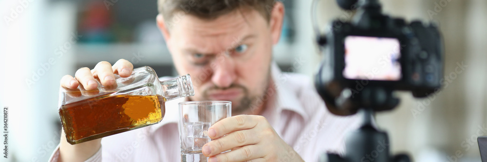 Portrait of man pouring whiskey into glass from bottle. Guy looking drunk and filming on videocamera. Unbutton shirt and bruise under eye. Production on camera Portrait of man pouring whiskey into glass from bottle. Guy looking drunk and filming on videocamera. Unbutton shirt and bruise under eye. Production on camera