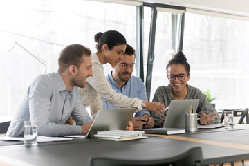 Smiling diverse young colleagues gather at desk in office look at laptop screen brainstorming at meeting together, happy multiracial employees cooperate using computer at briefing, teamwork concept