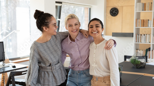 Portrait of smiling young multiracial female colleagues hug posing in modern office together, happy millennial diverse multiethnic women employees embrace at workplace, show friendship and unity