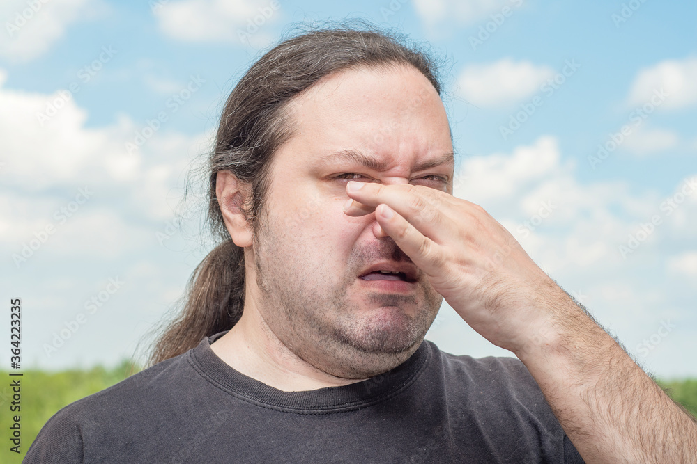 Obraz premium An ordinary middle-aged adult man with long tail hair pinches his nose with two fingers from a bad smell. Man on a light background.