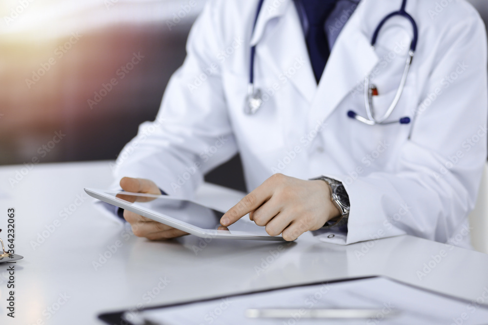 Unknown male doctor sitting and working with tablet computer iin a darkened clinic, glare of light on the background, close-up of hands Unknown male doctor sitting and working with tablet computer iin a darkened clinic, glare of light on the background, close-up of hands