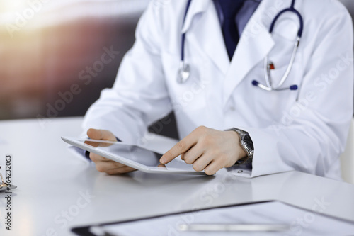 Unknown male doctor sitting and working with tablet computer iin a darkened clinic, glare of light on the background, close-up of hands