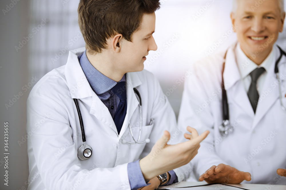 Doctors colleagues and elderly therapist discussing current disease therapy while sitting at working place in sunny clinic. Teamwork in medicine Doctors colleagues and elderly therapist discussing current disease therapy while sitting at working place in sunny clinic. Teamwork in medicine