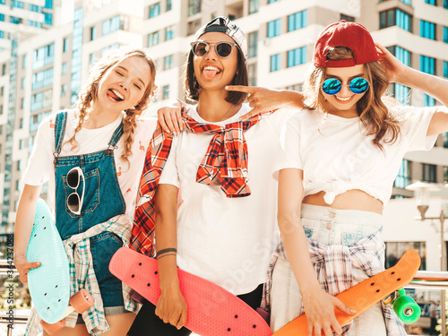 Three young smiling beautiful girls with colorful penny skateboards. Women in summer hipster clothes posing in the street background. Positive models having fun and going crazy