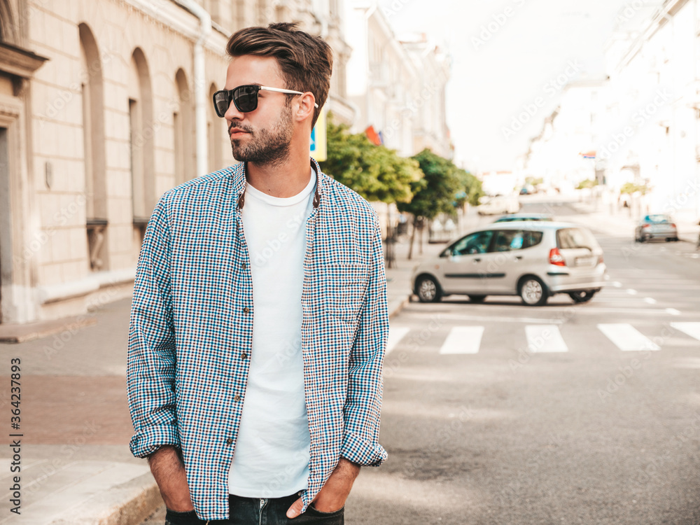 Fototapeta premium Portrait of handsome confident stylish hipster lambersexual model.Man dressed in white T-shirt. Fashion male posing on the street background in sunglasses
