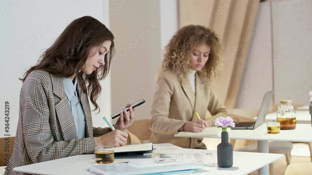 Medium shot of two young businesswomen sitting at desks in coworking space and writing in notebooks. Woman with dark hair recording voice message using smartphone Medium shot of two young businesswomen sitting at desks in coworking space and writing in notebooks. Woman with dark hair recording voice message using smartphone