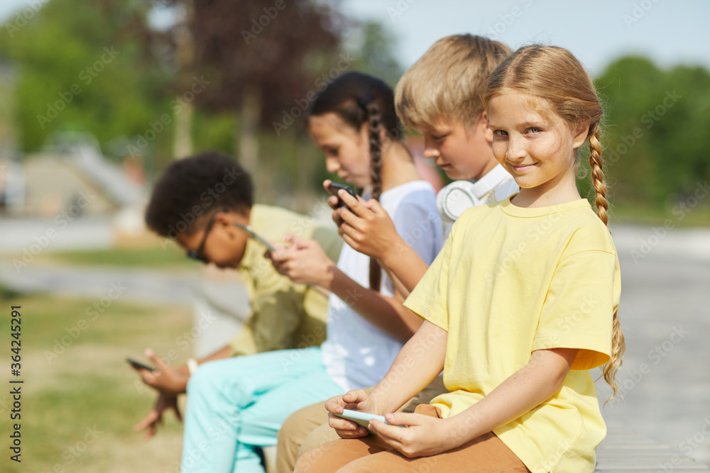 Multi-ethnic group of children using tablets and smartphones while sitting in row outdoors in sunlight, focus on smiling blonde girl in foreground, copy space Multi-ethnic group of children using tablets and smartphones while sitting in row outdoors in sunlight, focus on smiling blonde girl in foreground, copy space