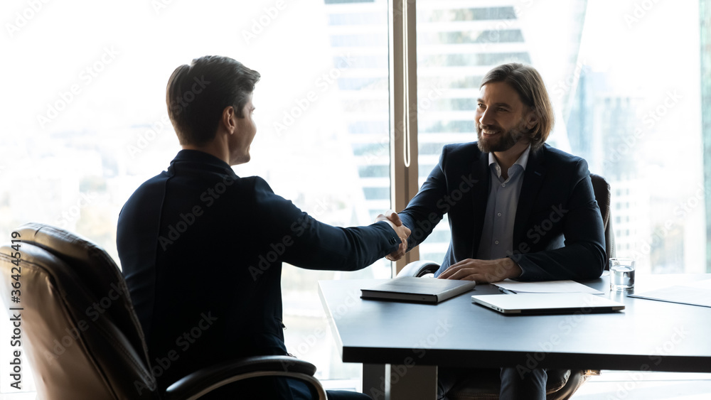 Happy two business partners shaking hands after negotiating cooperation details at meeting, smiling bearded professional financial advisor banker celebrating successful deal with client at office. Happy two business partners shaking hands after negotiating cooperation details at meeting, smiling bearded professional financial advisor banker celebrating successful deal with client at office.