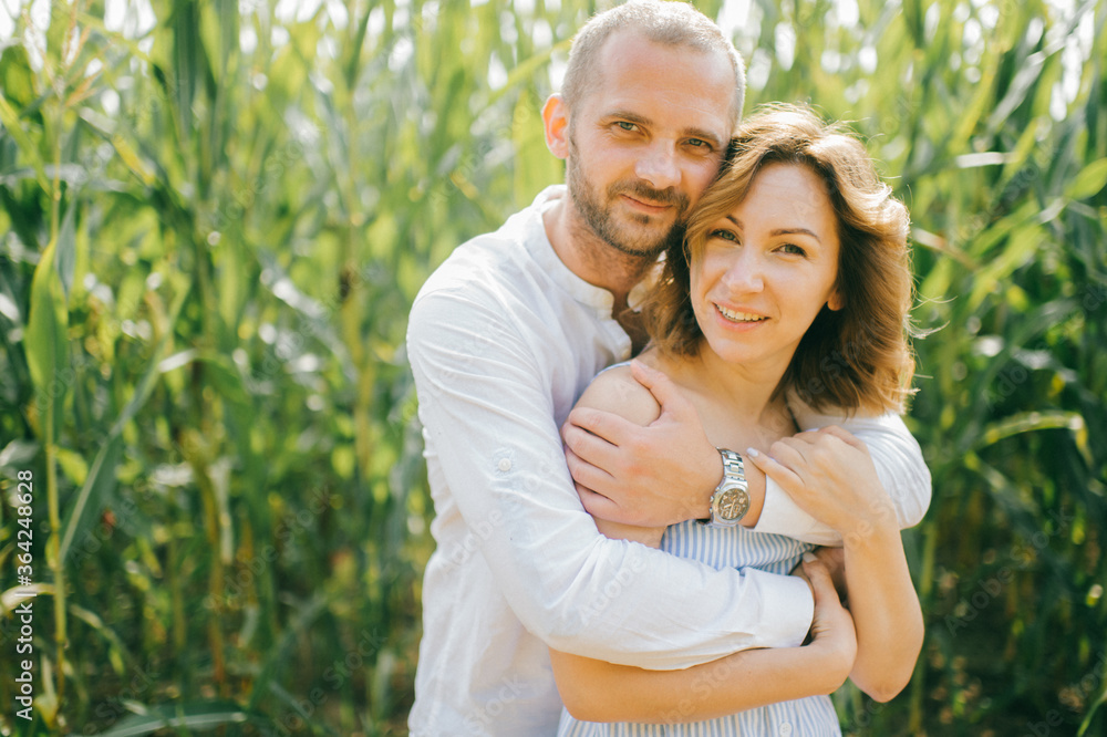 Attractive caucasian woman with short wavy dark hair in blue dress with her strong husband with short dark hair in white shirt and blue jeans relaxes together in the village cornfield Attractive caucasian woman with short wavy dark hair in blue dress with her strong husband with short dark hair in white shirt and blue jeans relaxes together in the village cornfield