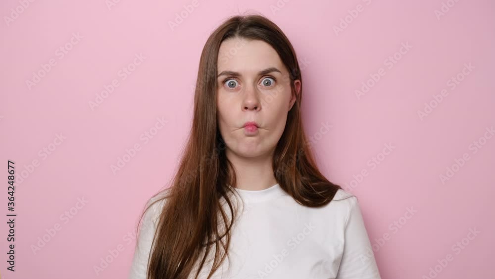 Surprised funny young woman poses indoor against pink studio background looking at camera with surprised big eyes, playful girl making fish face grimace pout lips, doing duck grimace, fooling around Surprised funny young woman poses indoor against pink studio background looking at camera with surprised big eyes, playful girl making fish face grimace pout lips, doing duck grimace, fooling around