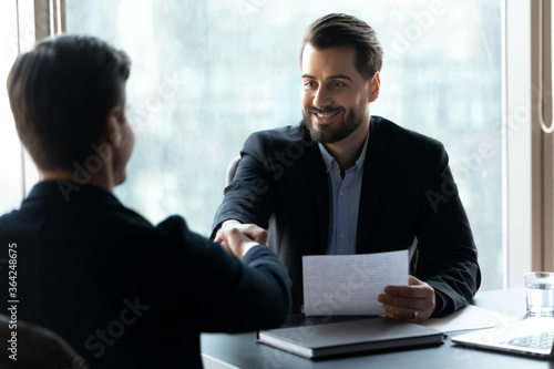 Smiling young handsome businessman in formal wear shaking hands with male confident partner after contract discussion at meeting, happy hr manager hiring promising job candidate at interview.