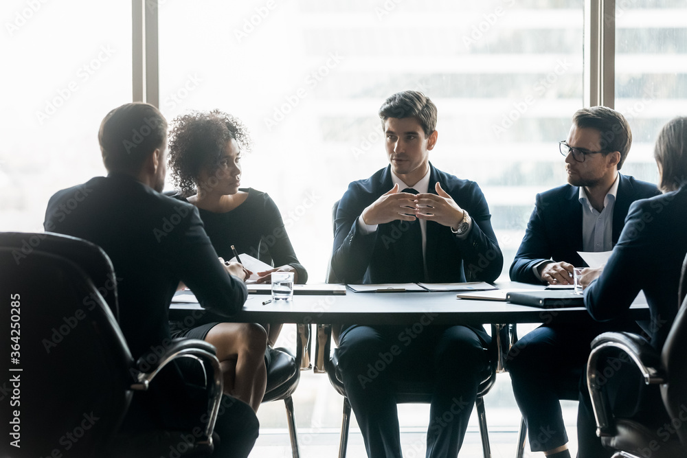 Focused young businessman holding negotiations meeting with mixed race colleagues in formal wear, sitting together ar desk in boardroom. Diverse ceo executive managers planning development strategy.