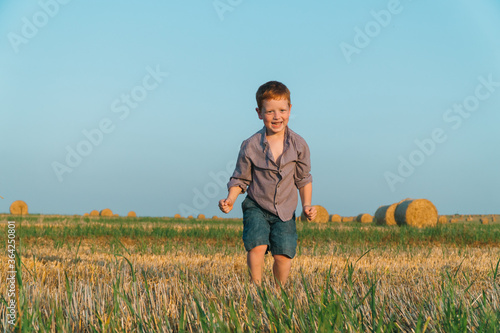 The red-haired boy goes ahead of a wheat field on a background of straw bales © voffka23