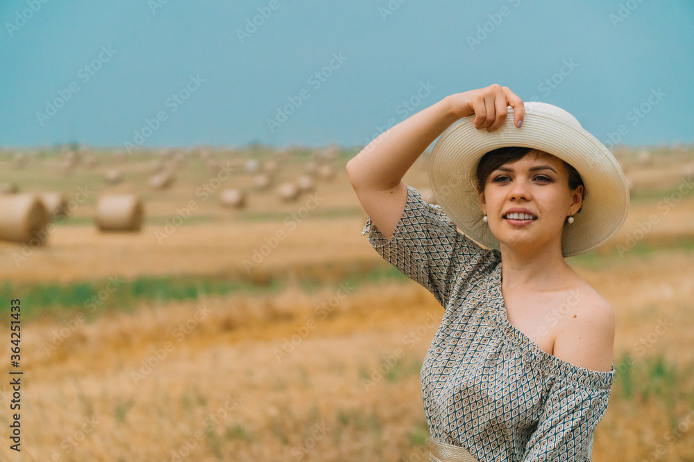 Beautiful girl in a hat looks into the sky in the middle of a wheat field with bales in summer evening