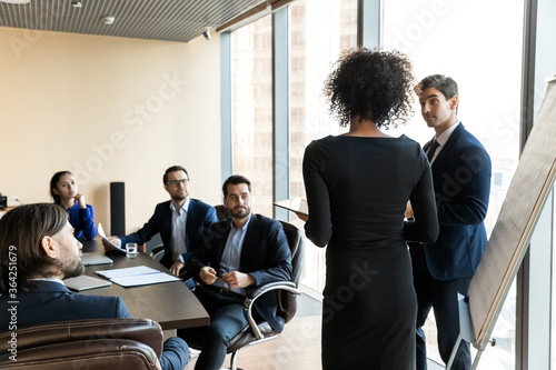 Back rear view young african american female speaker in formal wear standing near whiteboard with colleague, presenting project to interested investors partners clients or giving educational lecture.