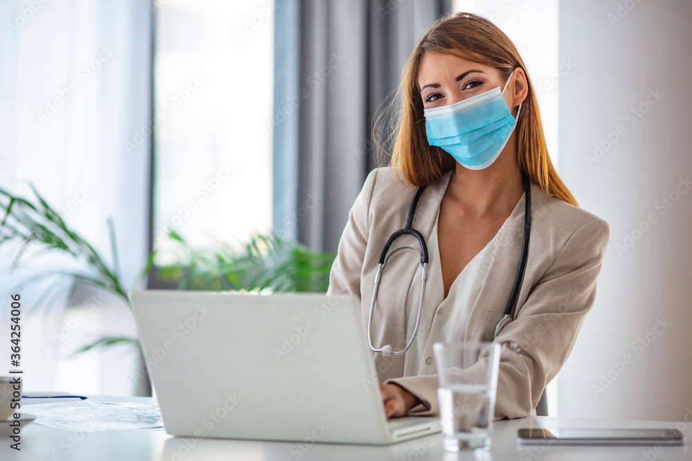 Female healthcare worker using laptop and looking at camera. Wearing protection mask during Corona Virus Outbreak. Doctor woman in protective medical mask sitting at table in office. Female healthcare worker using laptop and looking at camera. Wearing protection mask during Corona Virus Outbreak. Doctor woman in protective medical mask sitting at table in office.