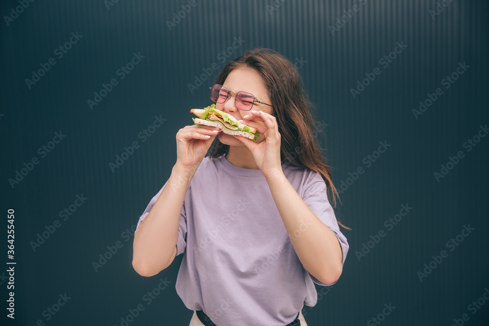 Young stylish trendy woman isolated over grey blue background. Hungry girl biting piece of fresh sandwich. Woman having breakfast or lunch outside at wall. Eating alone. Young stylish trendy woman isolated over grey blue background. Hungry girl biting piece of fresh sandwich. Woman having breakfast or lunch outside at wall. Eating alone.