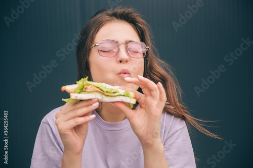 Young stylish trendy woman isolated over grey blue background. Girl enjoying eating tasty delicious sandwich during lunch or breakfast. Portrait of meal in hands.