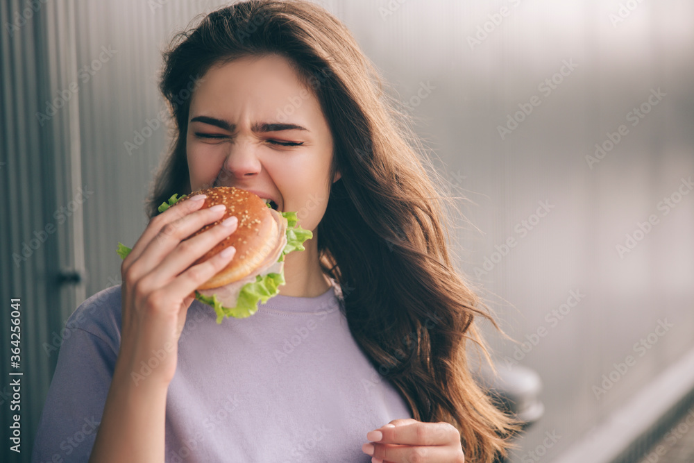 Young stylish trendy woman isolated over grey blue background. Hungry girl biting piece of burger. Lunchtime or dinner. Tasty delicious burger in hand. Young stylish trendy woman isolated over grey blue background. Hungry girl biting piece of burger. Lunchtime or dinner. Tasty delicious burger in hand.