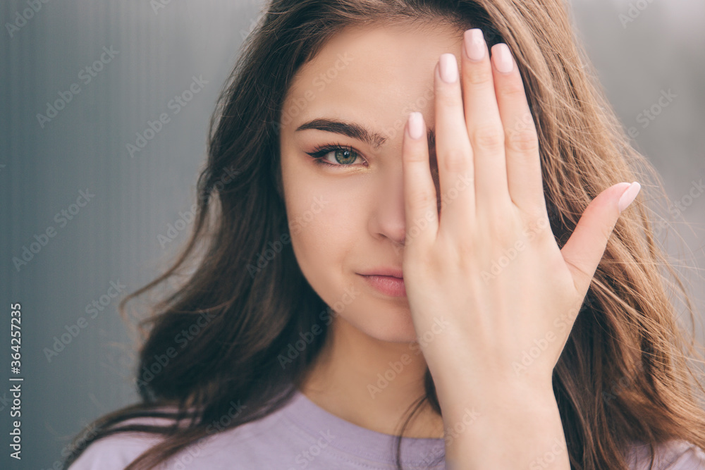 Young stylish trendy woman isolated over grey blue background. Beautiful portrait of amazing wonderful beautiful girl keeping one face part covered with hand. Looking straight forward.