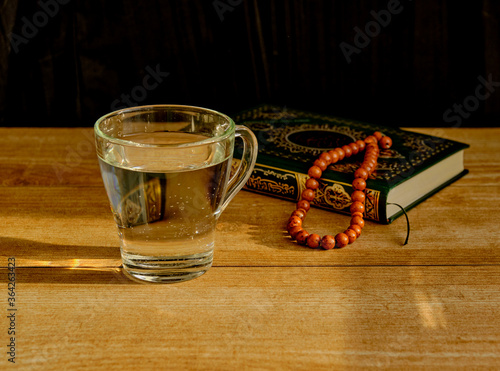 Glass of clear water, rosary and book of Quran on table during sunset. First drink, Muslim fast Eid al, Uraza photo