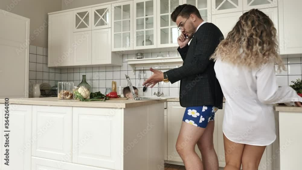 Serious busy business man talks on mobile phone discussing modern remote work opening laptop while little running son play game, wife do real housework. Family standing in comfortable kitchen together Serious busy business man talks on mobile phone discussing modern remote work opening laptop while little running son play game, wife do real housework. Family standing in comfortable kitchen together
