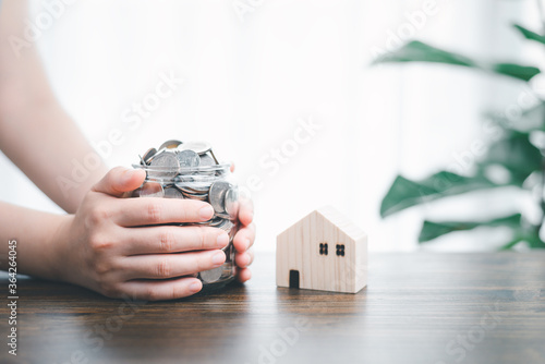 Two hands holding jar/bottle glass bank that contain full of coins at front is wood house model and tree house decoration background. Concept of financial money saving/ house or real estate investment