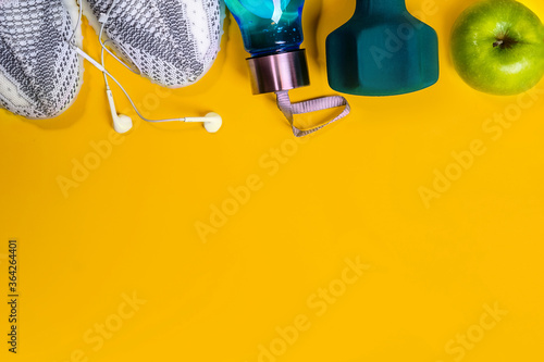 Fitness concept: dumbbell, trendy sport sneakers, bottle of water, headphones, apple, tennis ball on bright yellow background. Top view with copy space
