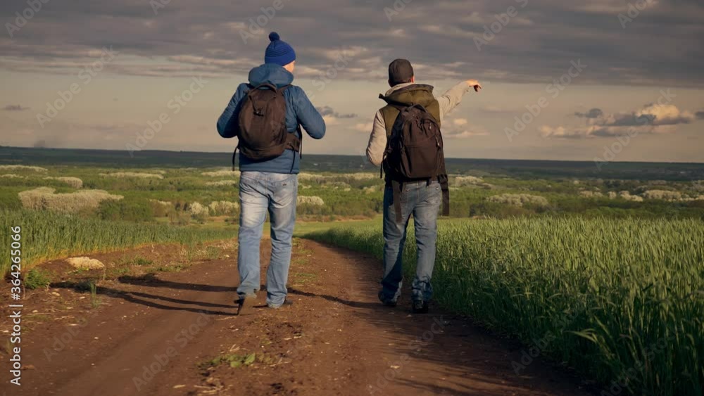 Team work. Two travelers with large foot backpacks walk down the road. Tourists lead an active lifestyle. Beautiful landscape of the field and sky. Team work. Two travelers with large foot backpacks walk down the road. Tourists lead an active lifestyle. Beautiful landscape of the field and sky.