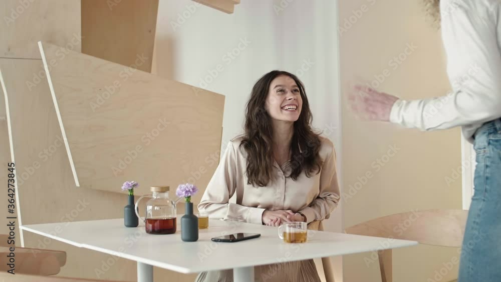 Medium shot of young pretty curly-haired woman is coming in cafe, greeting her cheerful female friend, sitting at cafe table and they are starting chat Medium shot of young pretty curly-haired woman is coming in cafe, greeting her cheerful female friend, sitting at cafe table and they are starting chat