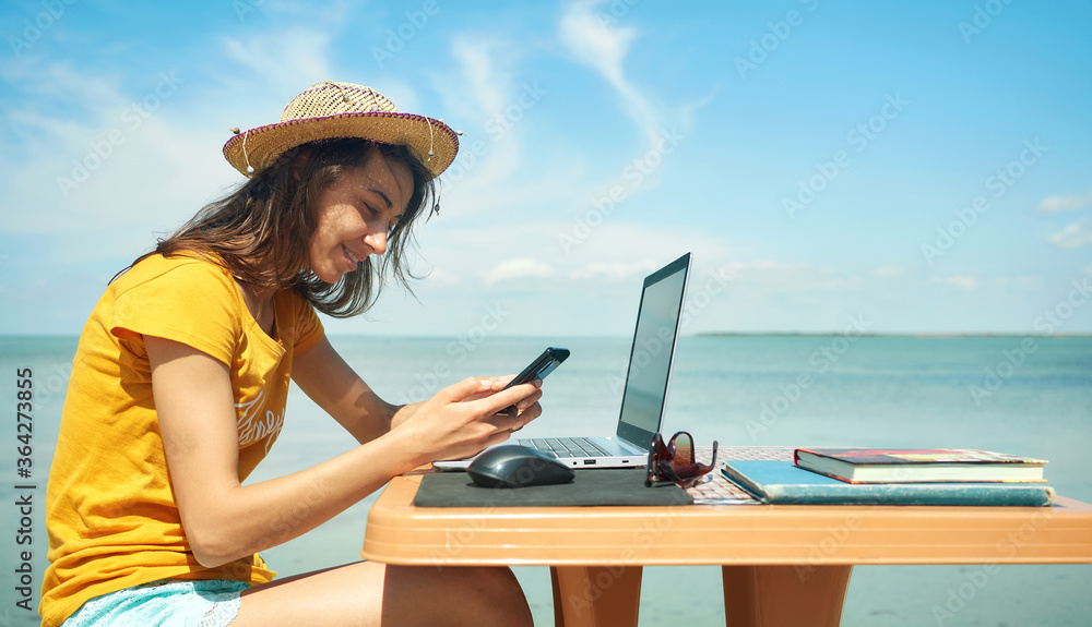 side view freelancer woman in hat with laptop computer working by sea at morning, using smartphone and internet. Technology and travel. Working outdoors. Freelance concept