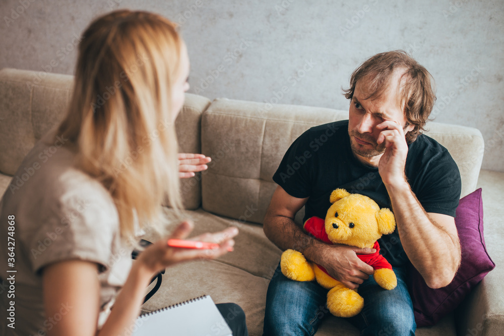 An adult male with psychological problems holds a teddy bear and cries in a psychologist office at a psychological session An adult male with psychological problems holds a teddy bear and cries in a psychologist office at a psychological session