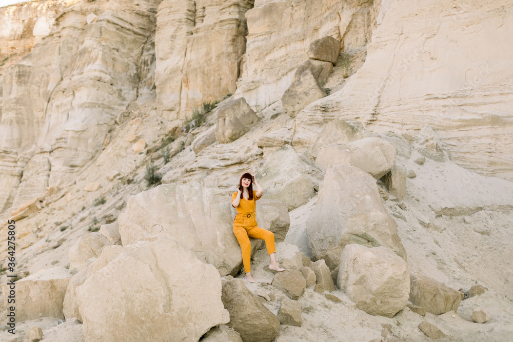 Summer outdoors portrait of young beautiful woman in fashionable yellow clothes, posing in sand quarry on sunny summer day, leaning on the sand rock. Pretty woman on the canyon cliff background