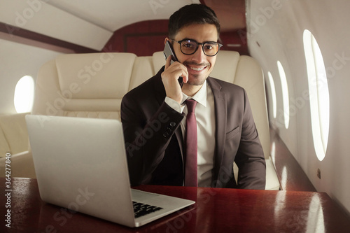 Handsome billionaire in his private jet, talking on phone with buisness partners, sitting at table with laptop ope © Damir Khabirov